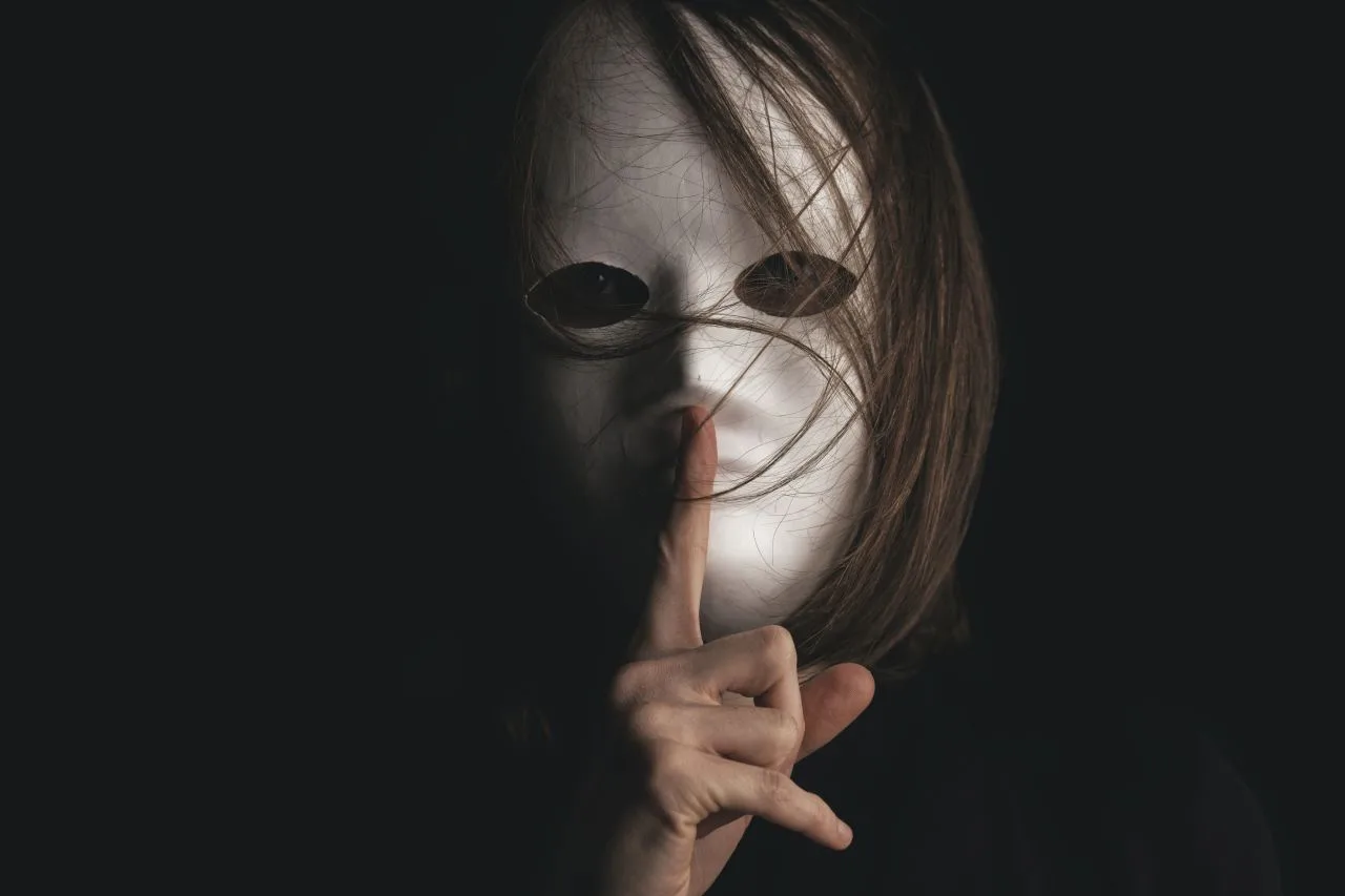 A person wearing a white mask holds a finger to the mask’s lips in a gesture of silence.