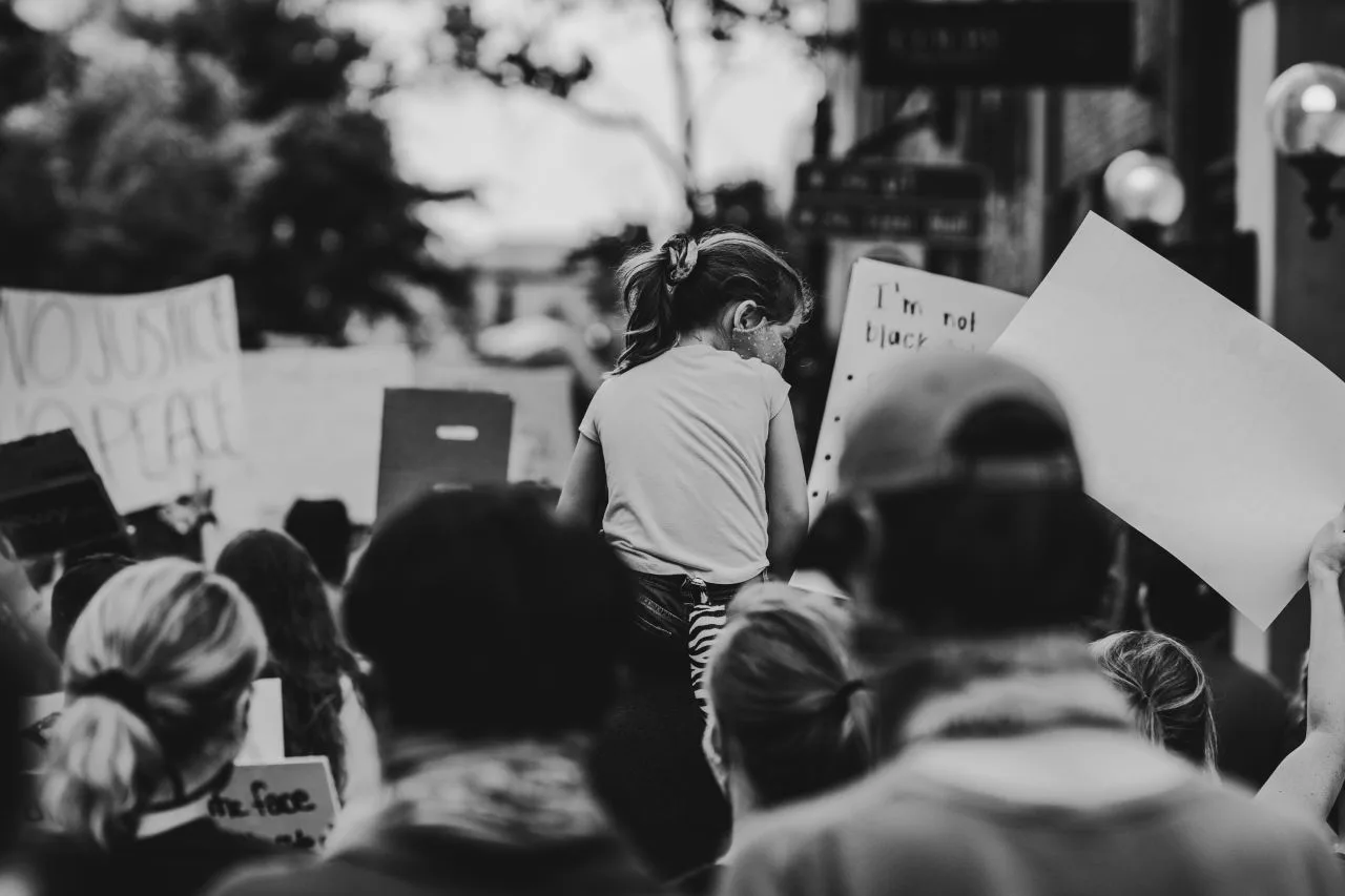 Black and white image of people marching with posters held up. Taken from behind. Centered, is a child sitting on someone's shoulders, towering over the other people marching.