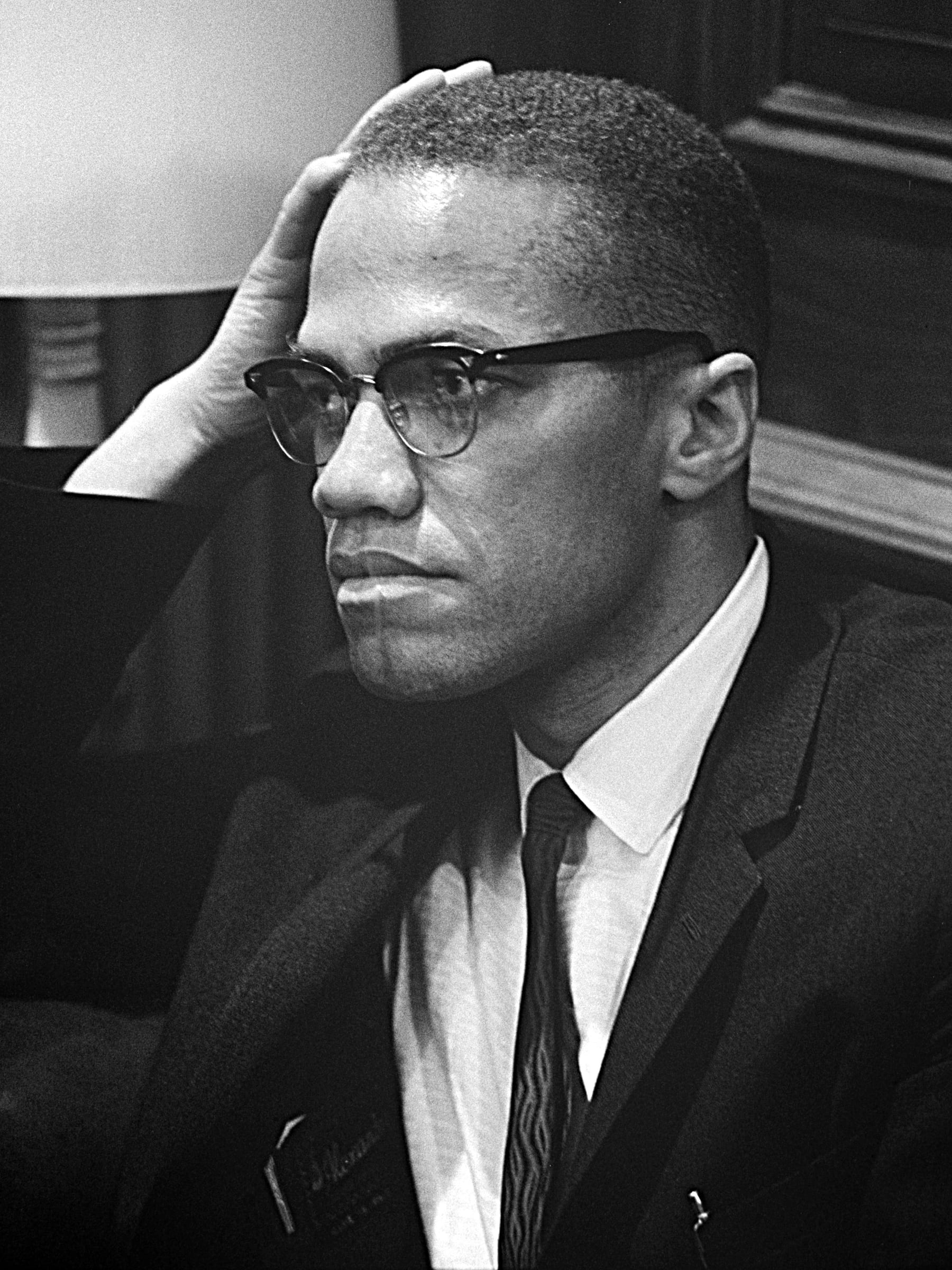 Seated at a press table in a suit and horn-rimmed glasses, resting his head against one hand.